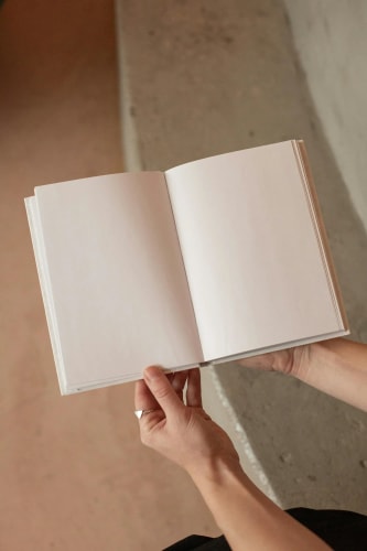 Une personne tenant un livre ouvert avec des pages blanches, en arrière-plan, un mur en béton et un sol en béton.