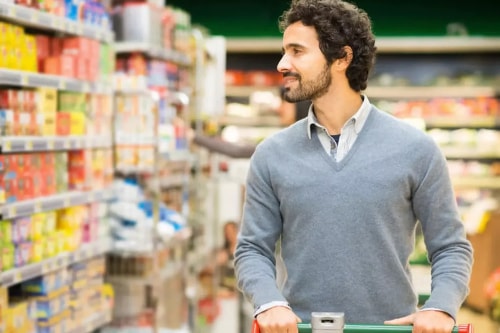 Un homme avec des cheveux bouclés et une barbe, poussant un chariot dans un supermarché, souriant en regardant les étagères remplies de produits, pour illustrer l'accompagnement sur l'offre par Pro Mundi Conseil