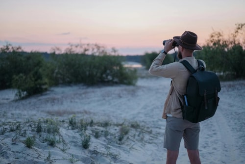 Un homme avec un chapeau et un sac à dos prend des photos d'un paysage naturel lors d'un coucher de soleil.