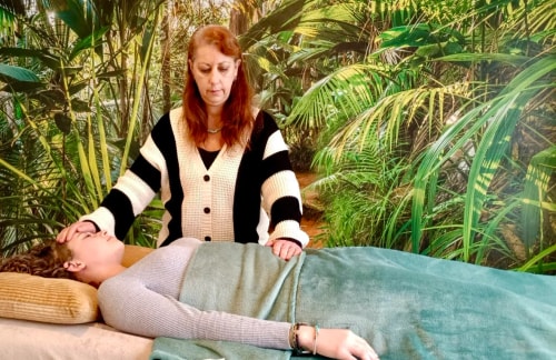 Corinne Duverger effectue un soin sur une autre femme allongée sur une table, entourée de plantes tropicales. La femme allongée est couverte d'une couverture verte.
