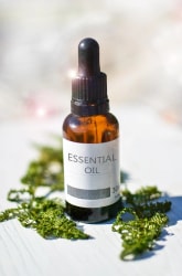 Flacon d'huile essentielle en verre ambré avec pipette, étiqueté "ESSENTIAL OIL", posé sur une surface claire entourée de feuilles vertes.