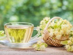 Tasse de tisane transparente avec des fleurs de tilleul et un panier de fleurs de tilleul sur une table en bois, flou de verdure en arrière-plan.