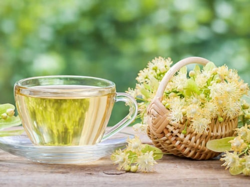 Tasse de tisane transparente avec des fleurs de tilleul et un panier de fleurs de tilleul sur une table en bois, flou de verdure en arrière-plan.