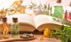 Un livre ouvert avec des bouteilles d'huiles essentielles et des gélules sur une cuillère en bois, entouré de fleurs et de plantes.