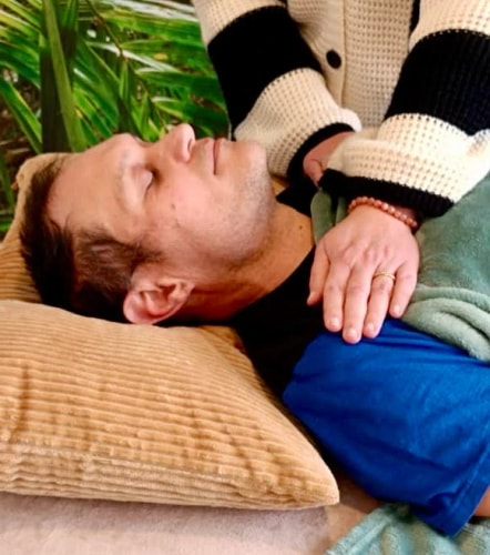 Un homme allongé avec les yeux fermés sur un coussin, tandis qu'une personne lui applique une technique de massage avec une main sur son épaule.