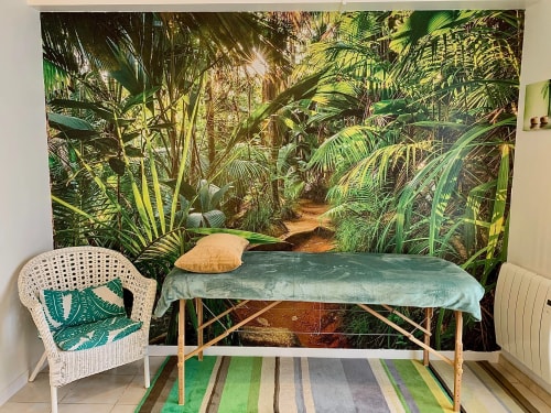 Bureau salle tropical avec un mur en arrière-plan représentant une jungle luxuriante, un lit de massage recouvert d'un drap vert et une chaise en osier à côté, sur un tapis rayé.