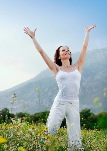 Femme souriante en vêtements blancs levant les bras au-dessus de sa tête dans un champ de fleurs avec des montagnes en arrière-plan.