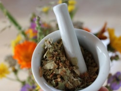 Un mortier avec des herbes séchées à l'intérieur, sur fond de fleurs colorées.