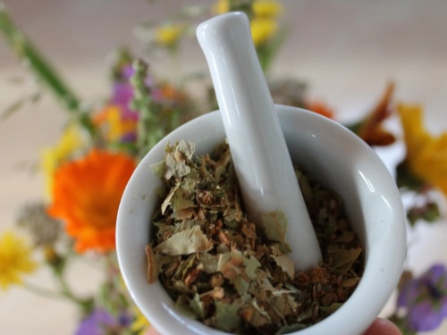 Un mortier avec des herbes séchées à l'intérieur, sur fond de fleurs colorées.