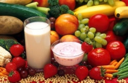 Un verre de lait à côté d'un bol de yaourt, entourés de divers fruits et légumes frais comme des fraises, des raisins, des carottes, des tomates, et des concombres.