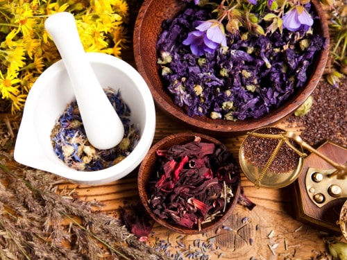 Un mortier et un pilon blancs à côté de bols en bois contenant des fleurs séchées multicolores, entourés de plantes et d'herbes sur une table en bois.