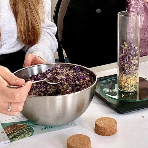 Une personne tient un bol en métal rempli de fleurs séchées colorées, pendant qu'une autre personne est visible en arrière-plan. Des ingrédients sont présentés sur une table.