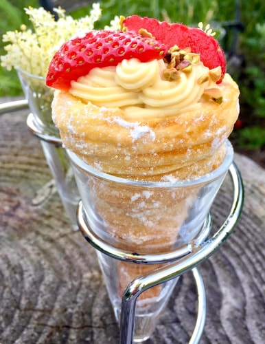 Un cornet parfumé à la crème de fleurs de sureau rempli de crème, garni de fraises et de morceaux de pistache, présenté dans un support en métal.