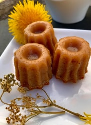 Trois cannelés dorés à la reine des près disposés sur une assiette blanche, accompagnés de fleurs jaunes et de brins de plantes séchées.