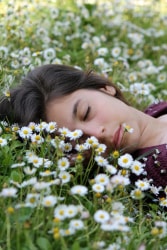 Une jeune femme allongée sur un tapis de fleurs blanches et jaunes, les yeux fermés, entourée de marguerites.