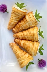 Cinq cornets apéritifs au fromage frais d'origanfeuilletés dorés, garnis d'une préparation crémeuse, disposés sur une assiette blanche avec des fleurs violettes et des feuilles vertes.