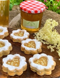 Confiture et biscuits au sureau noir en forme de fleur garnis de confiture sur une planche en bois, avec un pot de confiture rouge à carreaux et quelques fleurs d'ordinaire en arrière-plan.