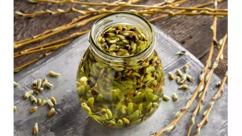 Un bocal en verre rempli de graines de cardamome, posé sur une surface en bois, entouré de tiges et graines éparpillées.