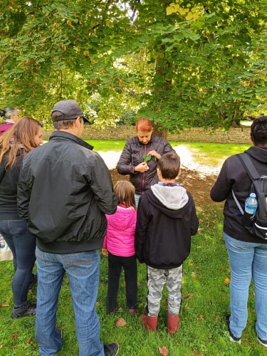 Un groupe de personnes debout sous un arbre, une femme montre quelque chose à des enfants.