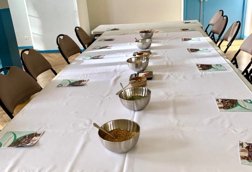 Vue d'une grande table dressée avec une nappe blanche, contenant plusieurs bols en acier inoxydable remplis d'épices, accompagnés de brochures sur le côté.