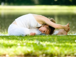Une personne pratiquant le yoga en posture de flexion avant, assise sur l'herbe près d'un plan d'eau.