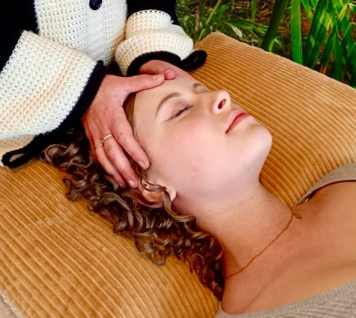 Une femme aux cheveux bouclés repose sur un coussin pendant qu'une autre personne lui tient le front, créant une atmosphère de détente et de bien-être.