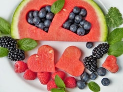 Une assiette de fruits composée de tranches de pastèque en forme de cœurs, accompagnées de bleuets, de mûres et de framboises, décorées de feuilles de menthe.