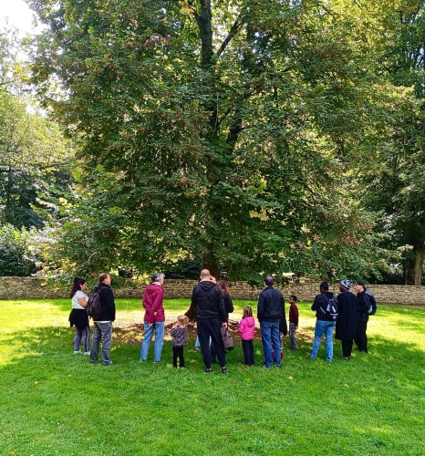 Un groupe de personnes, dont des enfants, se tient en cercle autour d'un grand arbre dans un parc verdoyant.