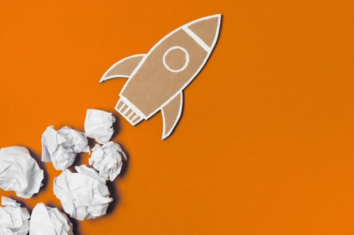 Une fusée en carton décollant d'un fond orange, entourée de boules de papier froissé blanc.