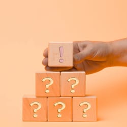 Une main tient un cube en bois marqué d'un point d'exclamation au-dessus de plusieurs cubes également en bois, marqués de points d'interrogation, sur un fond orange.