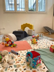 Espace de jeu pour enfants avec des tapis colorés, divers jouets en tissu et en plastique, et un petit module de motricité en mousse devant une grande fenêtre avec des barreaux.