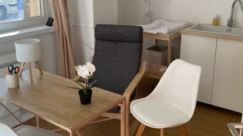 Chambre lumineuse avec lit aux couvertures superposées, coussin orange, plaid beige à franges et console blanche décorée de plantes.