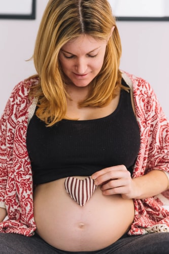 Femme enceinte blonde portant un débardeur noir et une veste à motifs, tenant délicatement un petit cœur en tissu rayé contre son ventre dénudé, avec un sourire bienveillant.