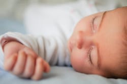 Une femme enceinte noire avec des dreadlocks, assise en tailleur contre un mur blanc, portant un haut noir, un gilet en jean sans manches et des bijoux, le regard tourné sur le côté en souriant.