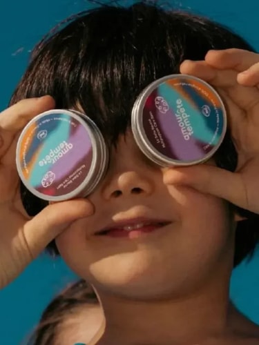 Un enfant aux cheveux bruns tient deux petits pots ronds devant ses yeux, imitant des lunettes, sur un fond bleu uni.