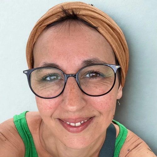 Portrait rapproché d’une femme souriante portant des lunettes rondes, un bandeau beige et un débardeur vert, sur fond clair.