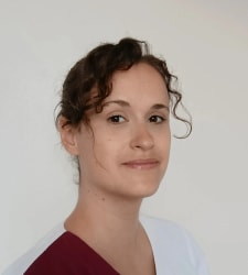 Portrait d’une jeune femme aux cheveux bruns bouclés, portant un haut bordeaux et blanc, sur un fond gris clair.