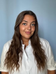 Portrait d’une jeune femme aux longs cheveux bruns ondulés, portant une blouse blanche, devant un mur bleu clair.