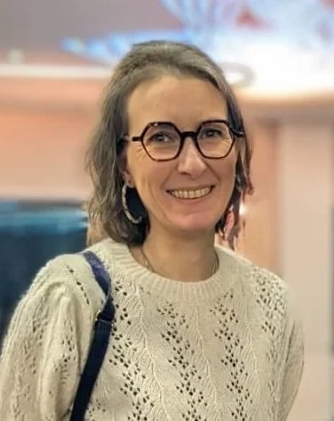 Portrait d’une femme souriante portant des lunettes noires et un pull blanc en maille, avec un sac à l’épaule, dans un intérieur lumineux.