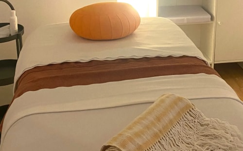 Chambre lumineuse avec lit aux couvertures superposées, coussin orange, plaid beige à franges et console blanche décorée de plantes.