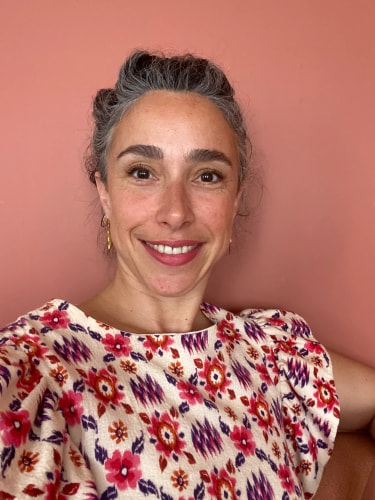 Portrait d'une femme souriante aux cheveux gris coiffés en chignon, portant un haut à motifs floraux colorés, devant un mur rose.