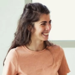 Femme souriante aux cheveux bruns mi-longs, vêtue d’un haut rose pêche, photographiée de profil sur un fond clair.