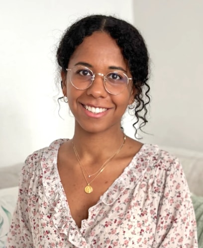 Portrait d'une jeune femme souriante, aux cheveux bruns bouclés, portant des lunettes rondes et un chemisier à fleurs. Elle porte un collier avec un pendentif doré. Fond blanc neutre.