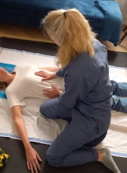 Une thérapeute aux cheveux blonds, agenouillée, pratique un soin énergétique ou une thérapie manuelle sur une personne allongée sur le dos, en posant doucement ses mains sur son abdomen et sa poitrine.
