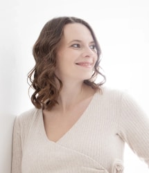 Portrait d’une femme aux cheveux bruns bouclés, vêtue d’un pull beige, souriant et regardant vers la droite sur fond clair.