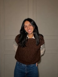 Jeune femme souriante aux longs cheveux noirs, portant un pull brun et un jean bleu, debout devant une porte grise.