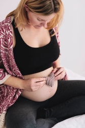 Femme enceinte souriante tenant un petit cœur en tissu rayé sur son ventre dénudé, portant un haut noir, un legging et un gilet à motifs.