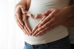 Les mains d'un couple forment un cœur sur le ventre d'une femme enceinte portant un t-shirt blanc et un jean.