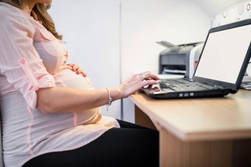 Une femme enceinte noire avec des dreadlocks, assise en tailleur contre un mur blanc, portant un haut noir, un gilet en jean sans manches et des bijoux, le regard tourné sur le côté en souriant.