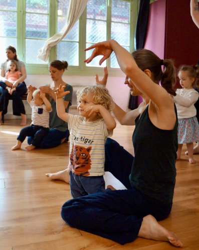 Une adulte et plusieurs enfants participent à une activité de danse ou de mouvement dans une salle lumineuse, les bras levés et en musique.